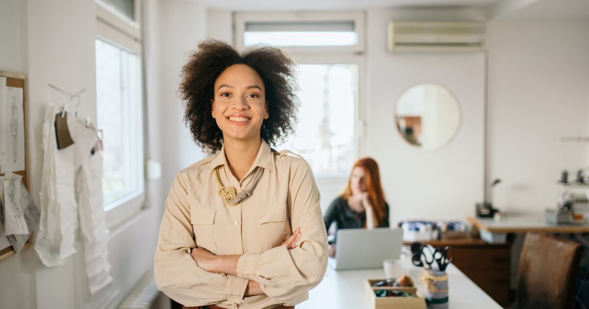 female leader confidently standing in conference room with staff person working in the background.